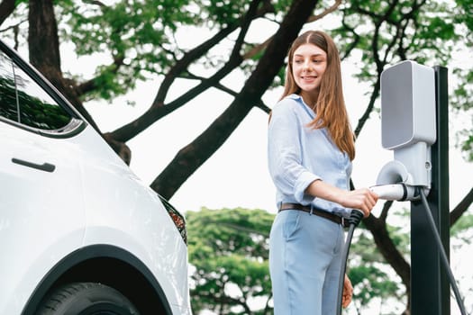 Young woman recharging battery for electric car during road trip travel EV car in natural forest or national park. Eco friendly travel during vacation and holiday. Exalt