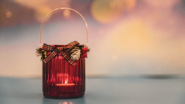 One red Christmas glass jar with a candle burning in it stands on the left on a blurred colorful background with bokke and copy space on the right, side view close-up.