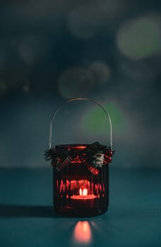 One red Christmas glass jar with a candle burning in it stands on a blurred dark background with bokeh, light reflection and copy space from above, side view close-up.