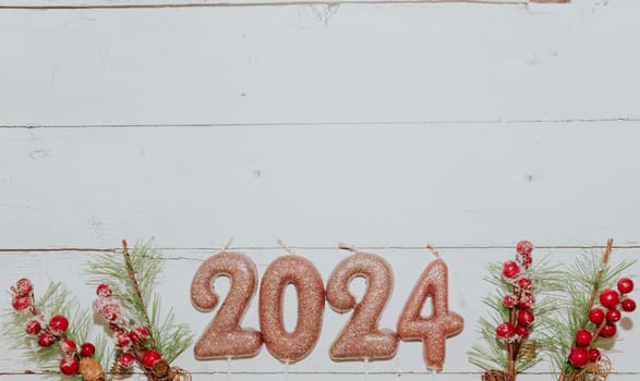 One festive light brown shiny candle number 2024 and Christmas fir branches lie below on a white wooden table with copy space on top, flat lay close-up.