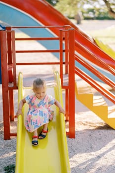 Little girl slides down a colorful slide at the playground, holding on to the railings. High quality photo