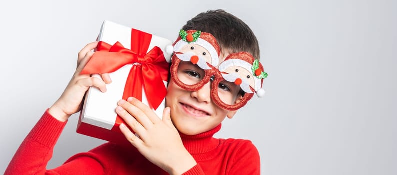 Smiling little boy wearing funny glasses in form of Christmas Santa Claus holding Xmas gift while standing in a white background, dressed in warm red xmas sweater.