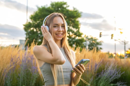 Optimistic blonde girl listens to the music via headphones standing in the park.