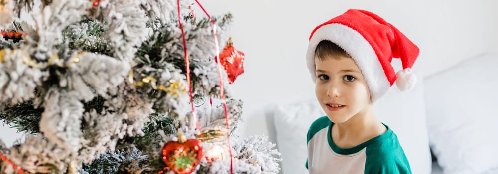 Portrait of cheerful boy smiling and laughing next to Christmas tree at house. Families and children generation alpha celebrating winter holidays.