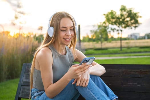Carefree blonde woman listening to music using headphones and select music via smart phone sitting on the bench in the park.