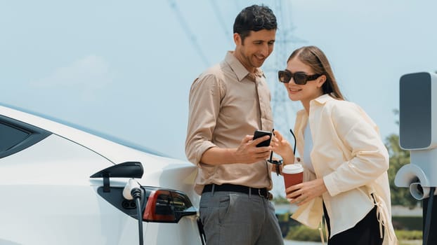 Young couple with coffee pay for electricity while recharge EV car battery at charging station connected to electrical power grid tower as electric industrial for eco friendly car travel. Expedient