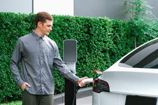 Young man travel with EV electric car charging in green sustainable city outdoor garden in summer shows urban sustainability lifestyle by green clean rechargeable energy of electric vehicle innards