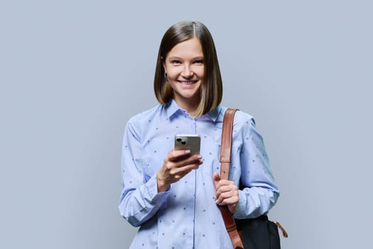 Young woman student with smartphone backpack on grey studio background. Smiling attractive female looking at camera. Using mobile applications apps for work business study leisure, technology, people