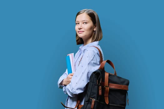 Portrait of young university student with backpack of textbooks on blue studio background. Smiling confident positive 20s female looking at camera. Education, knowledge, lifestyle, youth concept