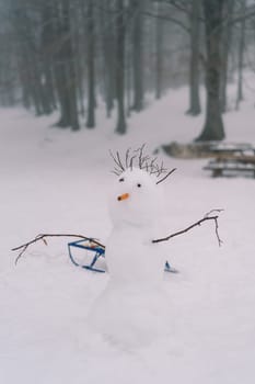 Snowman with stick hair and a carrot nose stands near a sled in a snowy meadow in the forest. High quality photo