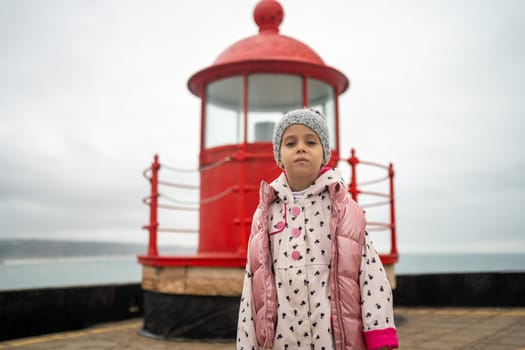 Little girl stands near red lighthouse on rocky seashore. Traveling and exploring seashore with red lighthouse in Nazare. Child dressed in warm clothes stands in frowning weather near lighthouse