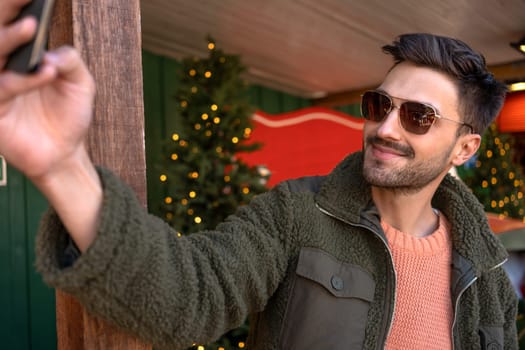 Man wear sunglasses taking selfie near Christmas store. Guy standing near Christmas tree smiling in winter season and taking picture of himself.
