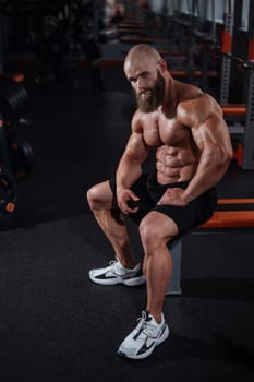 A muscular bald man in shorts is resting on a bench after a workout. Bodybuilder showing off his shape in the gym