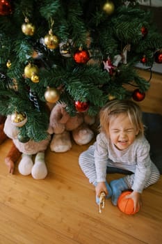 Small squinting girl with a ball in her hands sits on the floor near the Christmas tree. Top view. High quality photo