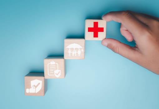 Health insurance and healthcare concept, human hand holds wooden block with icons about health insurance and healthcare access, retirement planning on blue background.