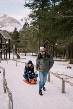 Smiling dad carries mom and little boy on a sleigh along a snowy path in the forest. High quality photo