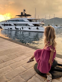 Little girl sits on a bollard on the pier and looks at a moored yacht at sunset. High quality photo
