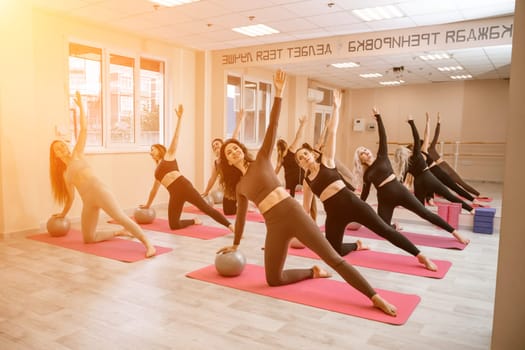 A group of six athletic women doing pilates or yoga on pink mats in front of a window in a beige loft studio interior. Teamwork, good mood and healthy lifestyle concept