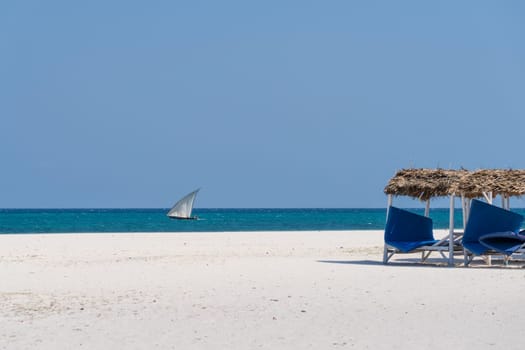 White sandy beach,turquoise ocean and deck chairs at the beach,background boat sails in the ocean at sunny day,luxury beach house.Zanzibar,Tanzania.