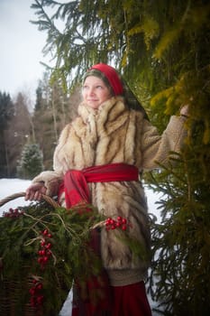 Teen girl in thick coat and a red sash with basket of fir branches and berries in cold winter day in a forest. Medieval peasant girl with firewood. Photoshoot in stile of Christmas fairy tale