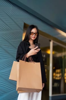 Woman using smartphone holding Black Friday shopping bag while standing on the side with the mall background.