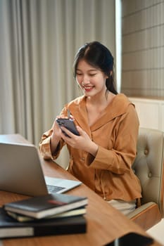 Charming asian female entrepreneur sitting in modern office and using mobile phone.