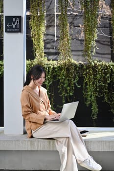 Portrait of happy young businesswoman working with laptop sitting outside her office building.
