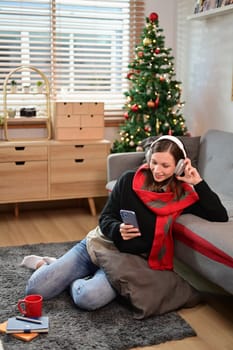 Smiling caucasian woman in headphone using mobile in living room decorated for Christmas.