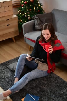Smiling young woman in warm clothes drinking tea and using digital tablet in living room.