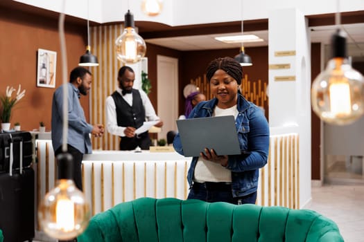 Happy millennial African American woman standing in hotel lobby looking at laptop screen, making video call while traveling alone. Female freelancer enjoying remote job, working remotely on vacation