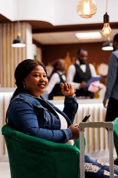 Happy smiling African American woman arriving at resort, sitting in hotel lobby with smartphone and waiting until check-in time. Black female guest sitting in lounge area using phone, traveling alone