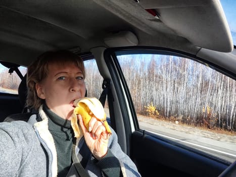 Portrait of female driver in solo journey. Adult mature middle aged woman holding steering wheel and a banana. Eating while driving for to stay awake. Lady girl with food while travel or trip by car