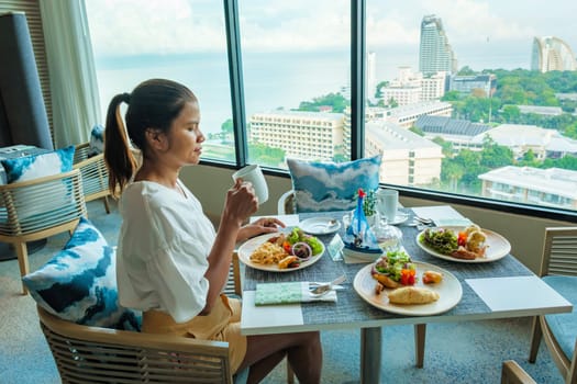 Asian Thai woman eating breakfast in a luxury hotel in Thailand, women drinking coffee looking out the window over the city and ocean