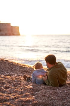 Dad is lying on the beach next to a little girl sitting and looking at the sea. Back view. High quality photo