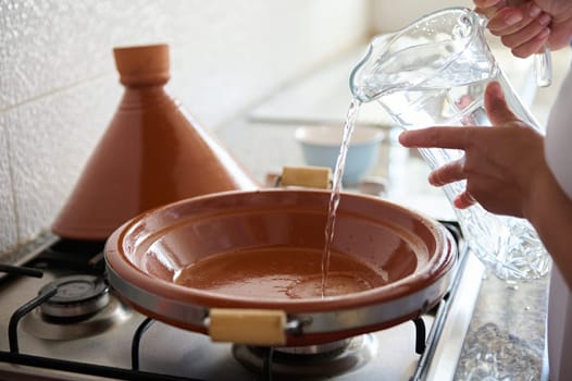 Closeup of an unrecognizable woman housewife pouring some water inside a tagine clay dish, cooking traditional Moroccan meal