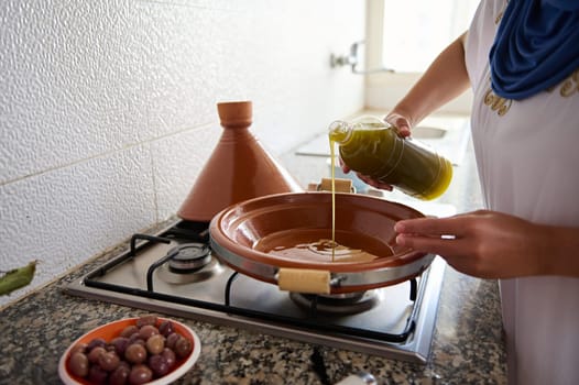 Closeup unrecognizable woman housewife beginning cooking meal in tajine pot, pouring extra virgin olive oil inside a clay dish standing on a stove. Traditional Moroccan food, culinary and culture