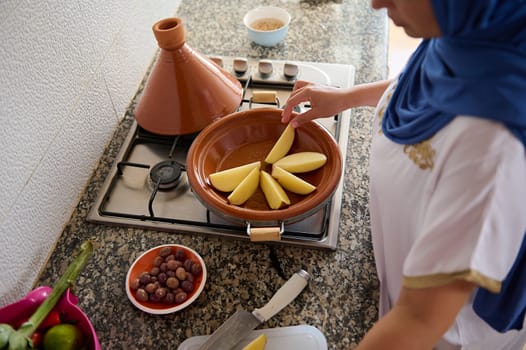 Top view Authentic Arab Muslim woman in hijab and long dress, standing by stove on kitchen counter and stacking potato slices in a clay pot, cooking Moroccan tajine for dinner in the kitchen at home