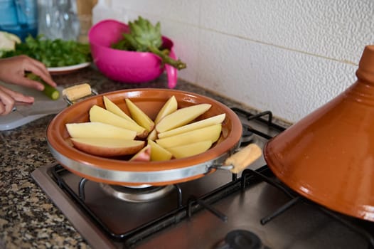 Sliced potato in clay dish on the stove on kitchen counter. Woman cooking tagine with fresh organic vegetables according to traditional Moroccan recipe. People. Culinary and national Oriental cuisine