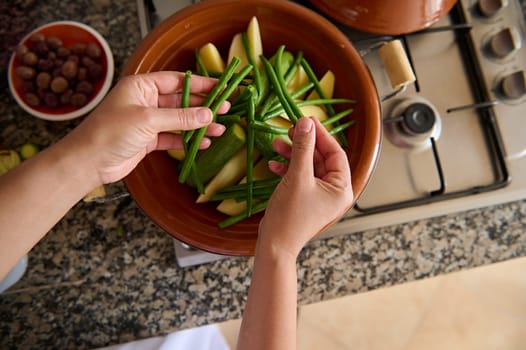 View from above of housewife hands putting fresh organic green beans on the tagine clay pot dish, while cooking dinner in the kitchen. Culinary. Cookery. Traditional Moroccan recipe and cuisine