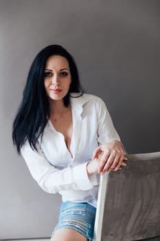 brunette woman in a white shirt sits on a chair on a gray background