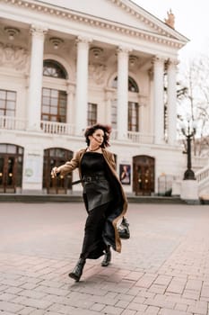 Woman street lifestyle. Image of stylish woman walking through European city on sunny day. Pretty woman with dark flowing hair, dressed in a beige raincoat and black, walks along the building