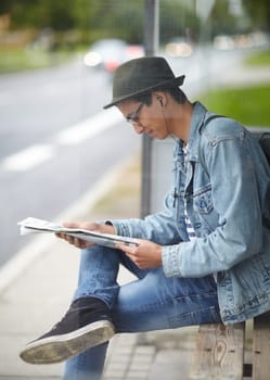 Man, commute and bus stop with newspaper, travel and adventure on journey, transportation and reading for trip. Male person, earphones and headlines or music, smile and happy for article in outdoors.