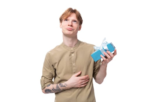 young red-haired man with glasses dressed in a brown shirt holds a blue box with a gift.