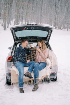 Couple drinking coffee from cups while sitting in the trunk of a car in a snowy forest. High quality photo