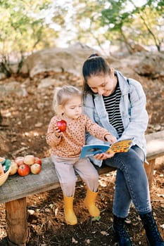 Little girl with an apple in her hand points to a book in her mother hands while sitting on a bench in the garden. High quality photo