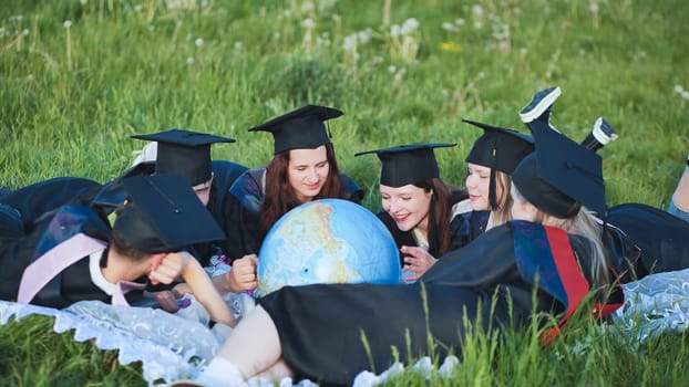 Graduates in black robes looking at a georgraphic globe lying on the grass