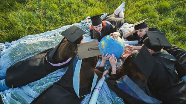 Graduates in black robes looking at a georgraphic globe lying on the grass