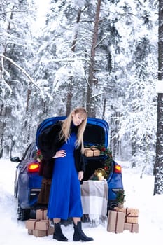 Woman in winter snowy forest in blue dress next to blue car decorated with Christmas decor. Christmas and winter holidays concept