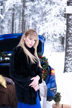 Woman in winter snowy forest in blue dress next to blue car decorated with Christmas decor. Christmas and winter holidays concept