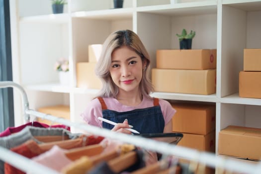 Portrait Of Asian Female Owner Of Fashion Store Checking Stock In Clothing Store With using notebook successful happy smile at small business, sme or ecommerce concepts.